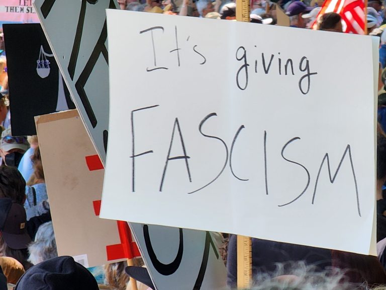 Surrounded by protesters and other signs, a sign on a white background says "It's giving FASCISM"