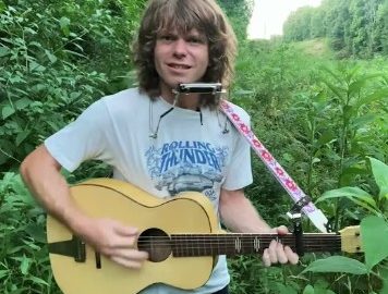 musician Jesse Welles stands in a field with a guitar and hands-free harmonica